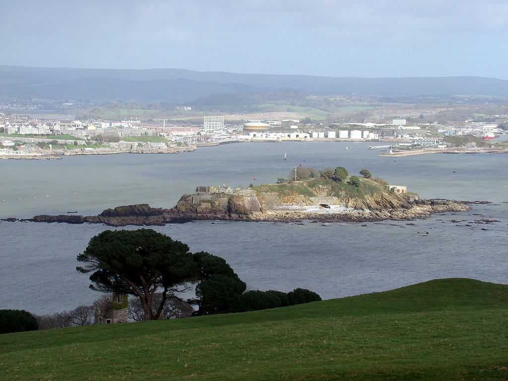 Northeastward view of Plymouth Sound from Mount Edgcumbe Country Park in Cornwall, with Drake's Island (centre) and, behind it from left to right, the Royal Citadel, the fuel tanks of Cattedown, and Mount Batten; in the background, the hills of Dartmoor.