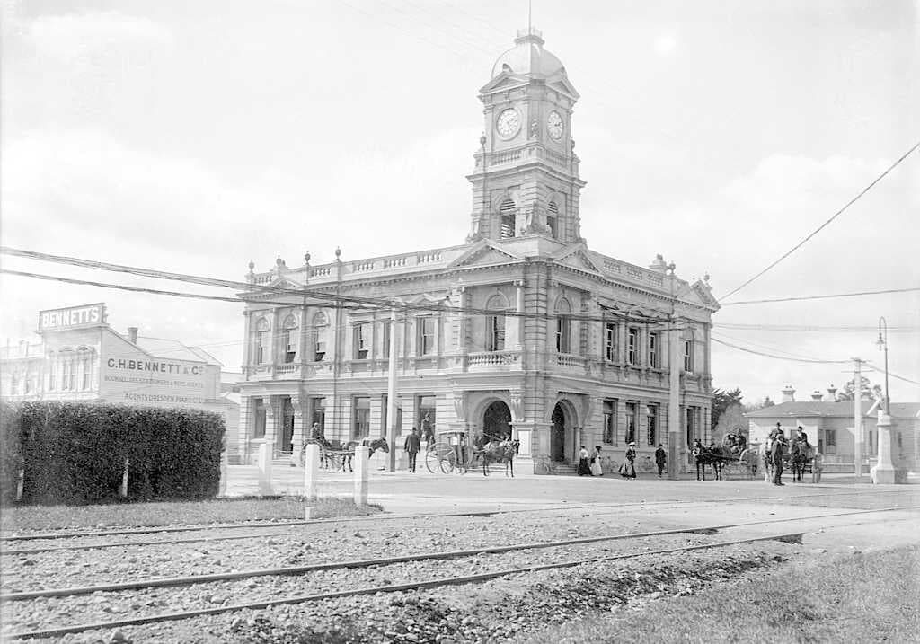 View of the (Chief) Post Office in Palmerston North, (opened in 1906) with the business premises of G H Bennett (booksellers, stationers & newsagents) behind a hedge centre left. There are a number of people, and horses & carriages outside the post office. Photograph taken by William A Price.